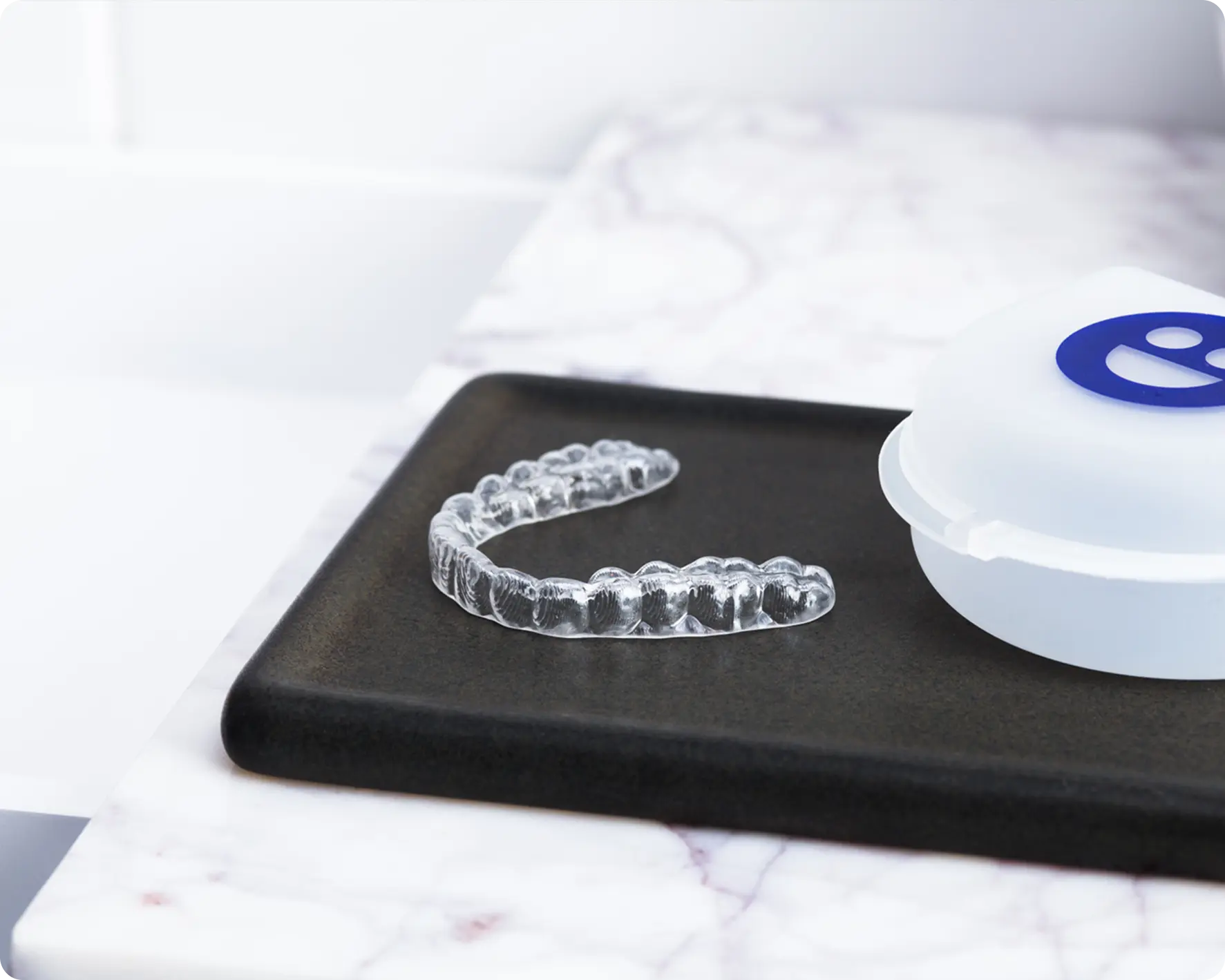 Clear dental aligner on a black tray with a white container featuring a blue logo.