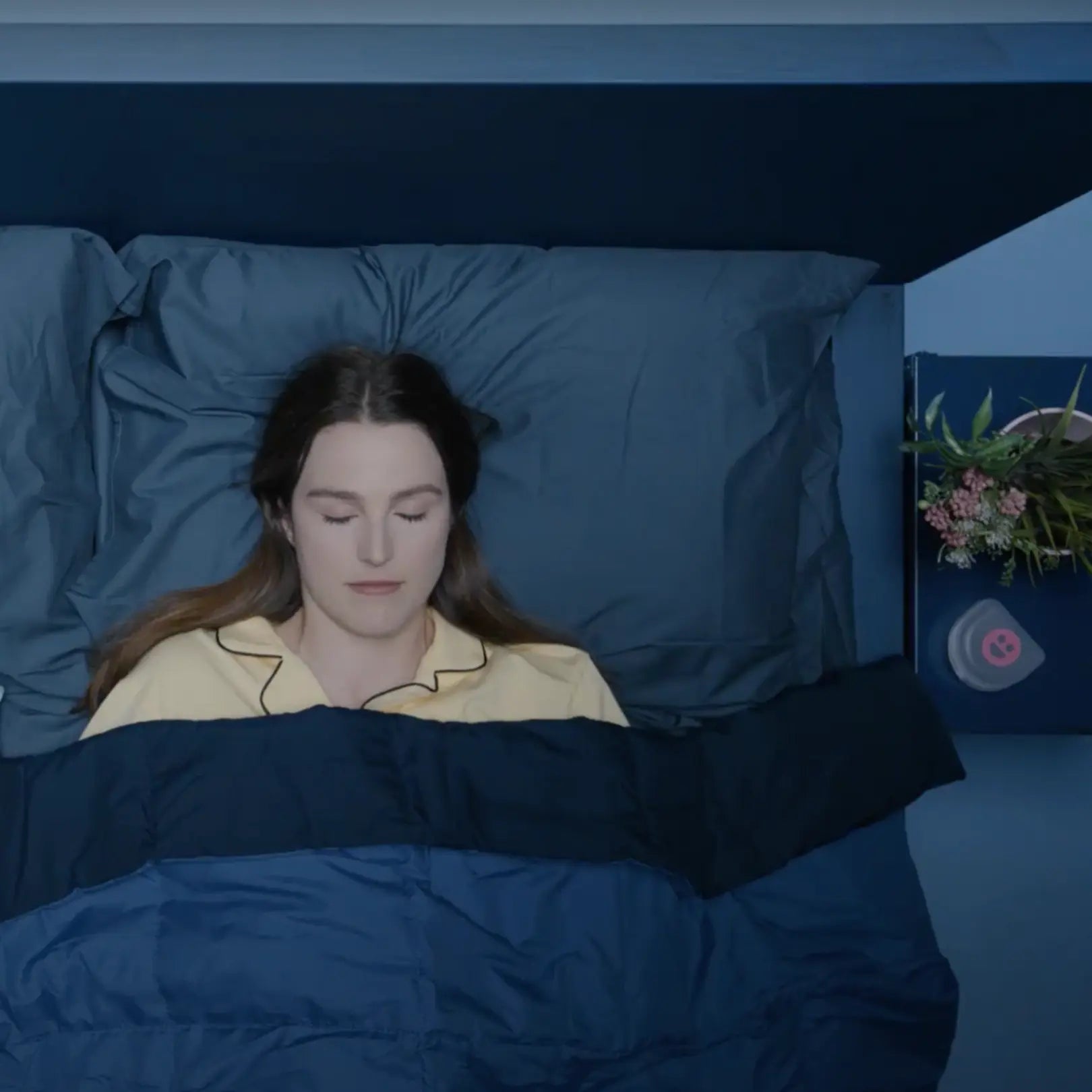 Woman lying in bed with blue bedding and a plant in the corner.