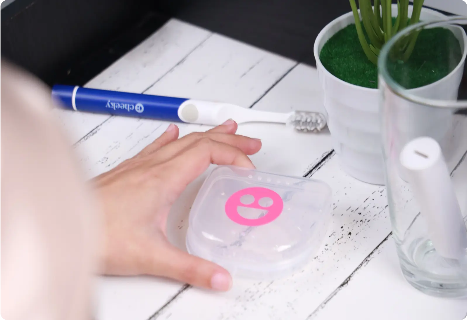 Hand reaching for a container with a pink logo on a desk with a pen, toothbrush, and plant in the background.