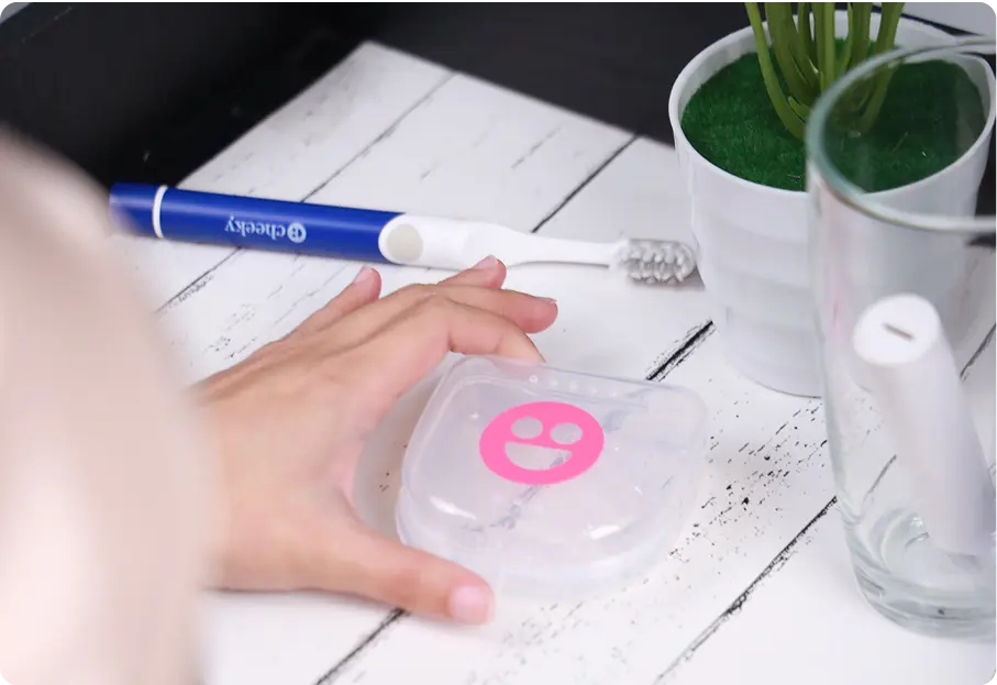 Person holding a clear container with a pink logo on a white surface with a toothbrush and pen in the background.