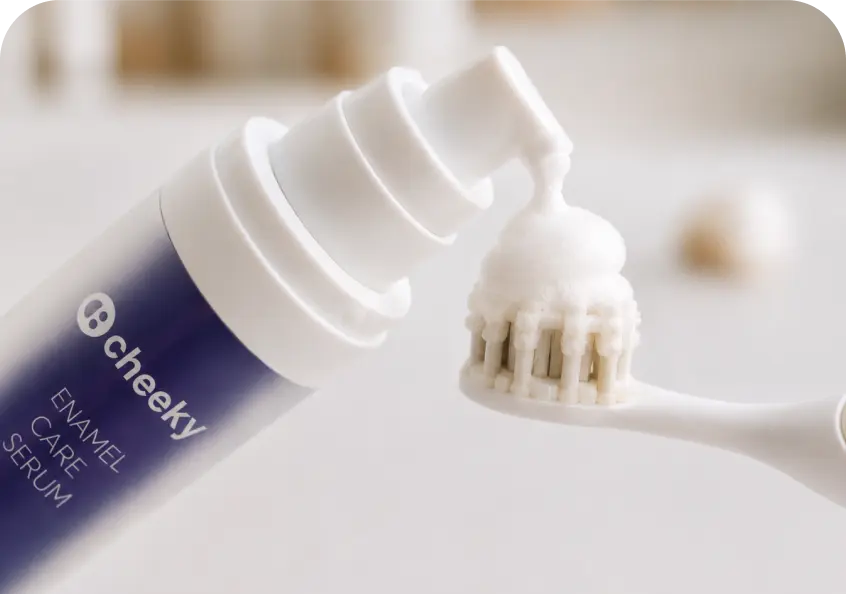 A close-up of a toothpaste tube labeled 'cheeky ENAMEL CARE SERUM' dispensing white foam onto a toothbrush head against a soft, blurred light background.