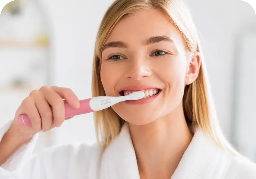 A smiling blonde woman in a white robe brushing her teeth with a pink electric toothbrush in a bright bathroom setting.